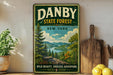 A vintage-style sign for the Danby State Forest in New York, featuring a scenic landscape with pine trees, a river, and mountains.