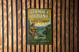 A metal sign with a vintage design hangs on a wooden wall, depicting a man in a historical outfit and the text "Daniel Boone Memorial State Forest Missouri: A Legacy of Adventure".