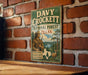 A vintage metal sign for Davy Crockett National Forest in Texas, featuring an illustration of a man in hunting gear and a deer in a forest setting.