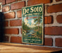 A vintage-style metal sign for De Soto National Forest in Mississippi, featuring a scenic landscape with a river, trees, and mountains.