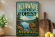A vintage metal sign for Delaware State Forest, featuring a scenic landscape with pine trees, a lake, and mountains.