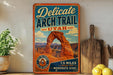 A vintage-style metal sign advertising the Delicate Arch Trail in Utah, featuring an illustration of the iconic rock formation.