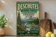 a vintage-style metal sign with a scenic mountain landscape and a river, placed on a wooden surface next to a wooden cutting board and some lemons.