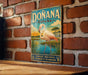 A vintage metal sign with a pink flamingo and a scenic landscape, mounted on a brick wall.
