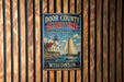 A vintage metal sign for Door County, Wisconsin, featuring a lighthouse and a sailboat on a lake.