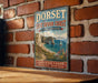A vintage-style sign for the Jurassic Coast in Dorset, England, featuring a scenic coastal landscape with cliffs, a beach, and a sailboat.