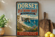 A vintage-style sign for the Dorset and East Devon Coast in the United Kingdom, featuring a scenic coastal landscape with cliffs, a beach, and sailboats.
