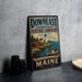 A vintage-style metal sign advertising Down East Coastal Heritage Landscape Maine, featuring a lighthouse and coastal scenery.