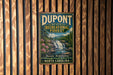 A metal sign with the text "DUPONT STATE RECREATIONAL FOREST" and a scenic image of a waterfall and forest, mounted on a wooden wall.