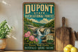A vintage-style sign for DuPont State Recreational Forest in North Carolina, featuring a scenic image of a waterfall surrounded by lush greenery and pink flowers.