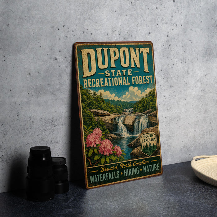 A vintage-style metal sign for DuPont State Recreational Forest, featuring a waterfall and lush greenery, is displayed on a gray concrete surface.