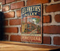 A vintage-style metal sign for Elfreth's Alley in Philadelphia, Pennsylvania, featuring an illustration of a brick street with a building and trees.