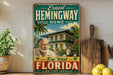 A vintage-style sign for Ernest Hemingway's home in Key West, Florida, featuring a painting of a man and a cat in a tropical setting.