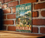 A vintage-style sign featuring a man and a cat, with the text "Ernest Hemingway Home" and "Key West Florida" printed on it, mounted on a brick wall.