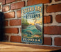 A vintage-style metal sign for Estero Bay Aquatic Preserve in Florida, featuring illustrations of various marine life such as a heron, a manatee, and a fish.