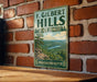 A vintage-style metal sign for F. Gilbert Hills State Forest in Massachusetts, featuring a scenic landscape with a river and pine trees.