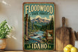 A vintage-style sign for the Floodwood State Forest in Idaho, featuring a scenic mountain landscape with a river flowing through it.