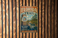 A vintage metal sign for the Florida Trail, featuring a scenic image of a trail surrounded by pine trees and a house, mounted on a wooden wall.