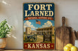 A vintage-style sign for the Fort Laredo National Historic Site in Kansas, featuring an American flag and a cannon, with a wooden paddle and a potted plant in the background.