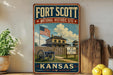 A vintage-style sign for the Fort Scott National Historic Site in Kansas, featuring an American flag and a wooden fence with a house in the background.