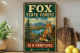 a vintage-style sign for the Fox State Forest in New Hampshire, depicting a fox sitting on a rock overlooking a forest and a lake.