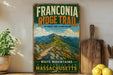 A vintage-style sign for the Franconia Ridge Trail in Massachusetts, featuring a scenic mountain landscape with a winding trail.
