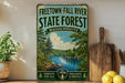 a vintage-style sign for the Freetown-Fall River State Forest in Massachusetts, with a scenic illustration of a river surrounded by trees and mountains. The sign is displayed on a shelf next to a wooden cutting board and a potted plant, creating a rustic and nature-inspired setting.