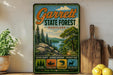 A vintage metal sign for Garrett State Forest, Maryland, featuring a scenic landscape with pine trees, a river, and mountains.