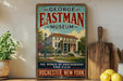 A vintage-style metal sign for the George Eastman Museum in Rochester, New York, featuring an illustration of the museum building surrounded by trees and flowers.
