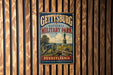 A vintage metal sign for Gettysburg National Military Park in Pennsylvania, featuring a cannon and a statue, is displayed on a wooden wall.