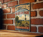 A vintage-style metal sign for Gettysburg National Military Park in Pennsylvania, featuring a cannon and a monument, is displayed on a brick wall.