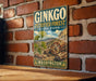 A vintage-style metal sign for Ginkgo Petrified Forest State Park in Washington, featuring a scenic illustration of a forest with ginkgo trees and a log cabin.