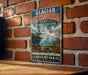 A vintage-style metal sign with a mountain landscape and text, mounted on a brick wall.