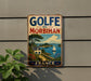 A vintage-style metal sign with the text "GOLFÉ DU MORBIHAN FRANCE" displayed on a wall, featuring a scenic landscape with a sailboat and a house.