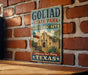 A vintage-style sign for Goliad State Park and Historic Site, featuring an illustration of a historic building and a wagon wheel.