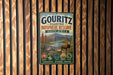 A metal sign with a vintage-style illustration of a landscape, featuring a gazelle, mountains, and a river, mounted on a wooden wall.