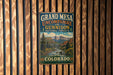 A vintage metal sign for Grand Mesa, Colorado, featuring a scenic mountain landscape with a river and pine trees.