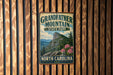 A vintage-style metal sign for Grandfather Mountain State Park in North Carolina, featuring a mountain landscape with pine trees and pink flowers.