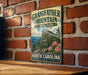 A vintage-style metal sign for Grandfather Mountain State Park in North Carolina, featuring a mountain landscape with pink flowers.