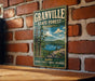 A vintage metal sign for Granville State Forest in Massachusetts, featuring a scenic image of a lake surrounded by pine trees and mountains.