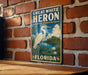 A vintage-style metal sign with a white heron and a tropical landscape, mounted on a brick wall.
