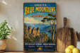 a vintage-style sign for the "Greater Blue Mountains Area of Australia", with a scenic image of the Blue Mountains in the background. The sign also includes the text "Spectacular Scenery - World Heritage" and "Explore, Discover, Experience", suggesting the area is a popular tourist destination.