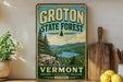A vintage-style sign for Groton State Forest, featuring a scenic mountain landscape with pine trees and a lake, is displayed on a shelf next to a wooden cutting board and some lemons.