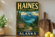 A vintage-style metal sign for Haines State Forest in Alaska, featuring a scenic mountain landscape with pine trees, a lake, and a road leading through the forest.