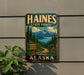 A vintage metal sign for Haines State Forest in Alaska, featuring a scenic mountain landscape with pine trees, a winding road, and a lake in the background.