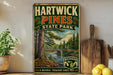 A vintage-style metal sign for Hartwick Pines State Park, featuring a scenic illustration of a forest and a river.