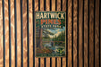 A vintage metal sign for Hartwick Pines State Park, featuring a scenic image of a forest and a river, mounted on a wooden wall.