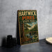 A vintage-style metal sign for Hartwick Pines State Park, featuring a scenic image of a forest and a river, is displayed on a gray concrete surface.