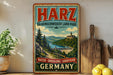 A vintage-style metal sign for Harz, Braunschweiger Land Park, featuring a scenic landscape with a castle, mountains, and a river.