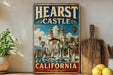 A vintage-style metal sign with the text "HEARST CASTLE CALIFORNIA" and an illustration of a castle surrounded by palm trees and a fountain.
